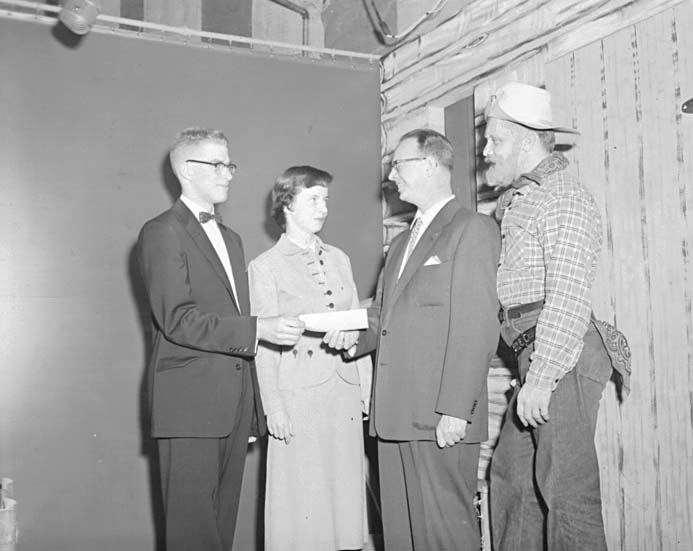 A presentation in the studio: two men in suits and a woman handing over an envelope, with Ted Knight in his Windy Knight western costume standing at the right.