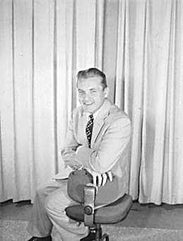 Publicity portrait of Ted Knight seated on a stool in a light suit, smiling, against a curtain backdrop.