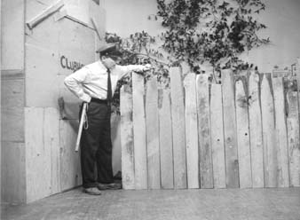 Ted Knight in a police uniform with cap and nightstick, leaning around the end of the clubhouse fence.