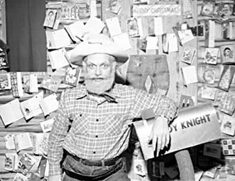 Ted Knight in a cowboy hat and plaid shirt as Windy Knight, standing in front of a wall covered with viewers' photographs and holding a box marked with his name.