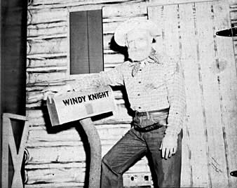 Ted Knight in western costume leaning against a sign reading WINDY KNIGHT on a log cabin set.