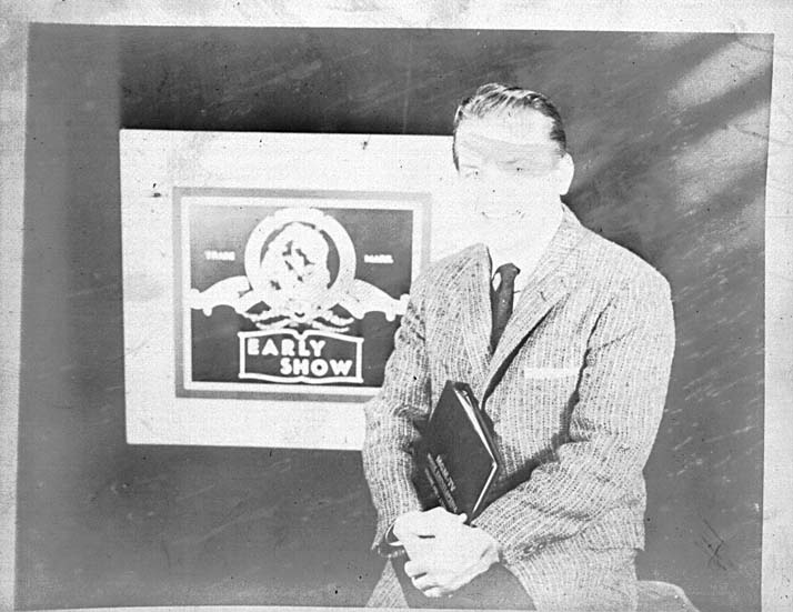 Ted Knight in a suit holding a folder, standing beside a painted title card reading EARLY SHOW.