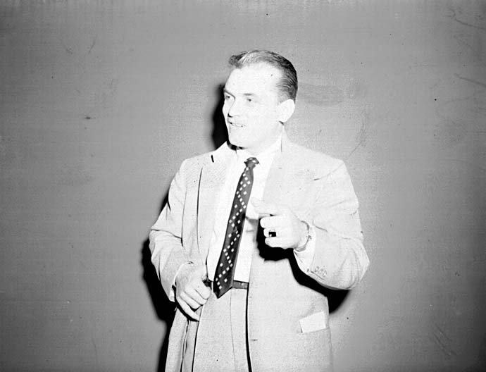 Ted Knight in a light suit and patterned tie, caught mid-gesture as if speaking to camera.