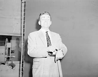 Ted Knight standing in a suit beside studio equipment, holding a hat.
