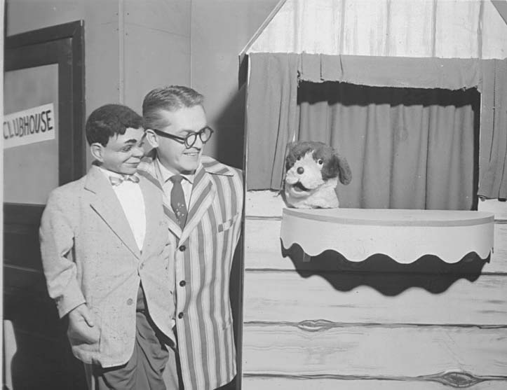 Ted Knight in a striped jacket and round glasses beside a ventriloquist's dummy, next to a door marked CLUBHOUSE, with a dog puppet in the window.