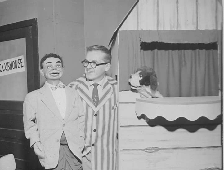 Ted Knight smiling toward the dog puppet in the clubhouse window, the dummy at his side.