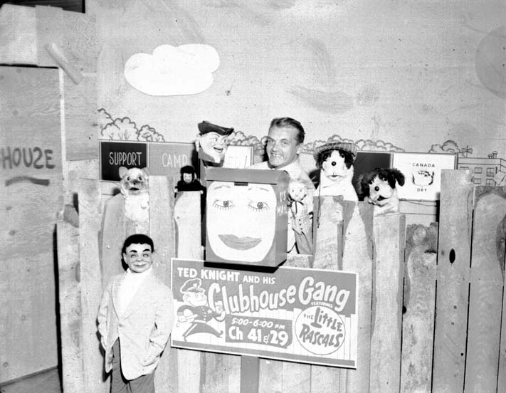 Ted Knight in a peaked cap behind a wooden fence lined with puppets, above a hand-painted sign reading "Ted Knight and his Clubhouse Gang, featuring The Little Rascals."