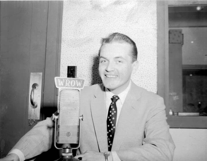 Ted Knight in a light suit smiling behind a ribbon microphone marked WROW in a radio studio.
