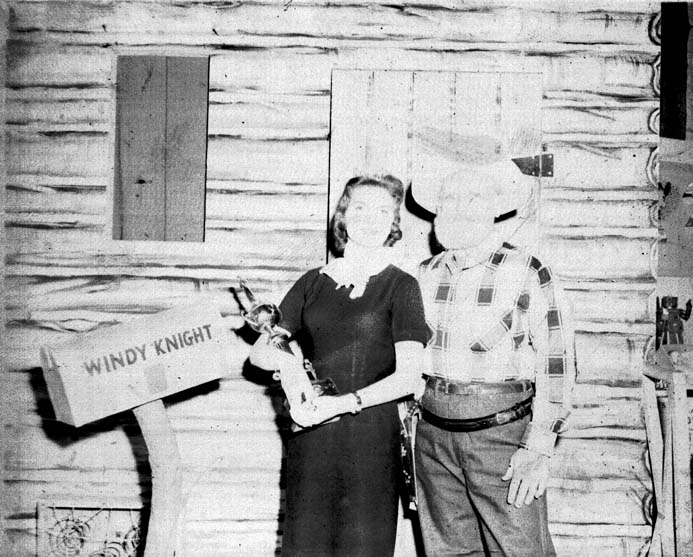 Windy Knight standing beside a young woman holding a trophy, in front of the log cabin set and the WINDY KNIGHT sign.