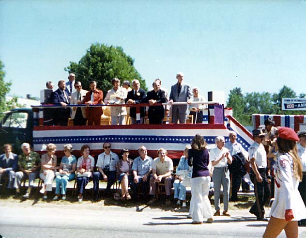 The parade seen from the crowd, the convertible passing along a tree-lined street.